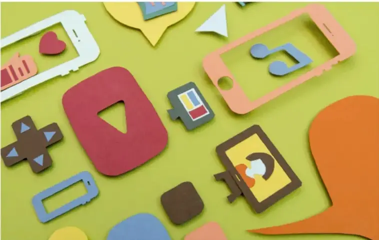 Recorte fotográfico de papel de colores con siluetas de dispositivos tecnológicos: teléfonos, una tableta con nota musical, burbujas de diálogo, íconos de corazón, video, cámara, control de videojuego y una tarjeta de crédito, sobre fondo verde, representando la cultura digital.