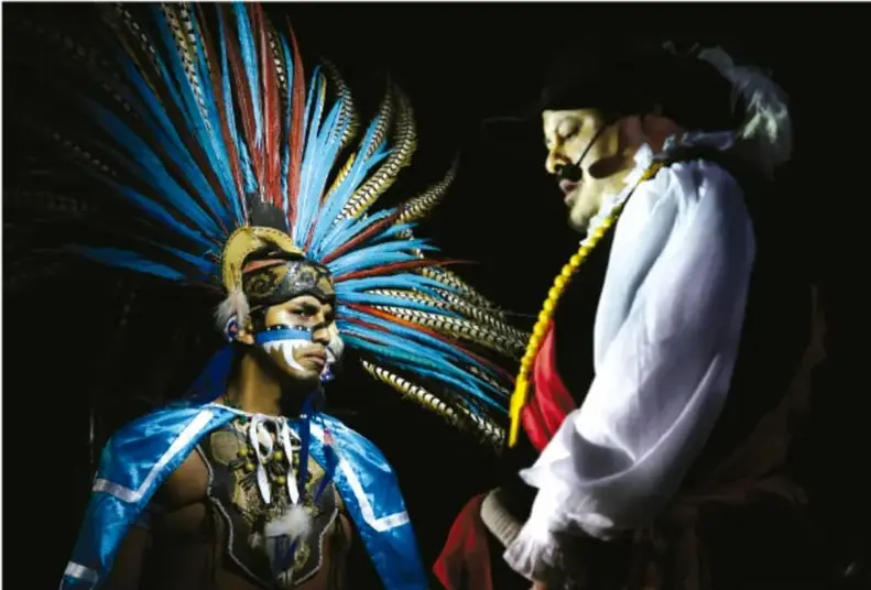 Fotografía de una escenificación teatral: a la izquierda un danzante con penacho azul y rojo y pintura facial; a la derecha un actor con máscara blanca, camisa blanca y listón rojo, sobre fondo negro.