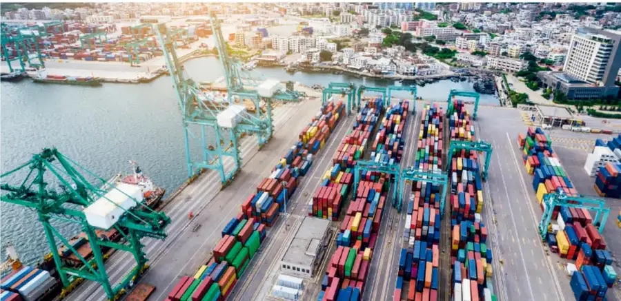 Fotografía aérea de un puerto marítimo con grúas pórtico turquesa, contenedores de carga de colores apilados y una ciudad costera al fondo; ilustra actividades de transporte y comercio internacional (sector terciario/secundario).