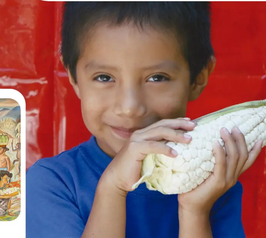 Fotografía de un niño sonriente, de camiseta azul, que sostiene con ambas manos un elote blanco grande frente a un fondo rojo.