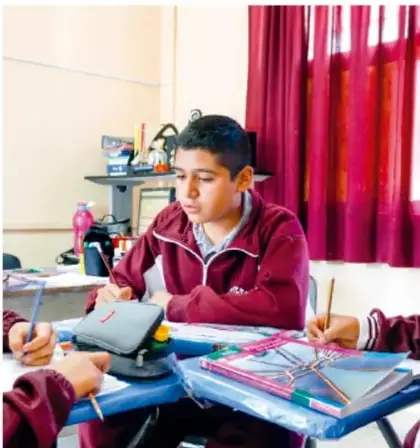Estudiantes de telesecundaria trabajando sentados en su escritorio con cuadernos y materiales escolares, en un aula con cortinas rojas.