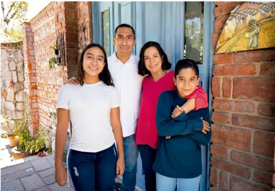 Fotografía de una familia de cuatro integrantes (dos adultos y dos adolescentes) sonriendo frente a una puerta azul, junto a una pared de ladrillo con una maceta y un cuadro decorativo.