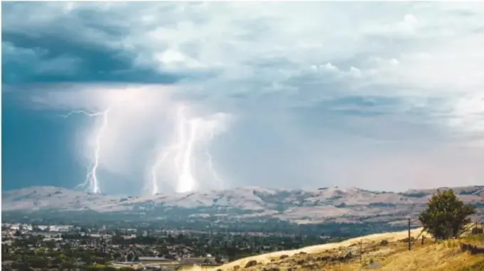 Fotografía de varios rayos ramificados cayendo de nubes de tormenta sobre un valle con cerros secos.