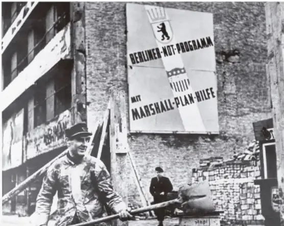 Fotografía histórica de un soldado frente a un cartel que dice 'BERLINER NOT-PROGRAMM' y 'MARSHALL-PLAN-HILFE', en un edificio en ruinas.