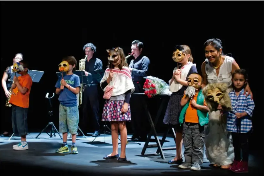 Fotografía de un grupo de niñas, niños y adultos sobre un escenario oscuro. Los niños llevan máscaras de animales (zorro, búho, lobo) y algunos adultos tocan saxofón y trombón. Al centro hay un atril con un ramo de flores rojas. Representa la presentación final de un proyecto artístico escolar.