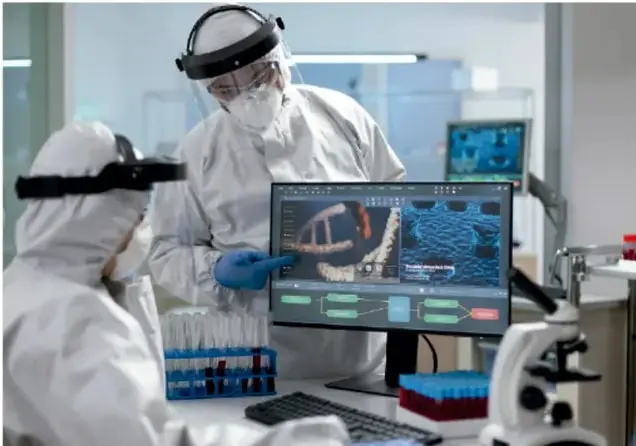 Fotografía de dos personas con trajes blancos de protección, mascarillas y caretas trabajando en un laboratorio frente a una computadora que muestra imágenes científicas; hay tubos de ensayo y un microscopio sobre la mesa.