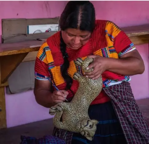 Fotografía de una mujer indígena de rasgos y vestimenta tradicional colorida (huipil), sentada y trabajando artesanalmente con un objeto tejido, en un entorno doméstico sencillo; ejemplifica la conservación de la identidad y las costumbres propias.