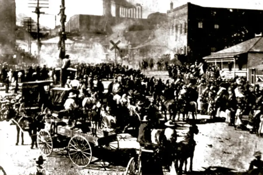 Fotografía histórica en blanco y negro de un día de multitud en las calles de Cananea, con obreros a pie, jinetes, coches de mulas, postes de luz y edificios de la compañía minera al fondo, durante la huelga de ca. 1906-1907.