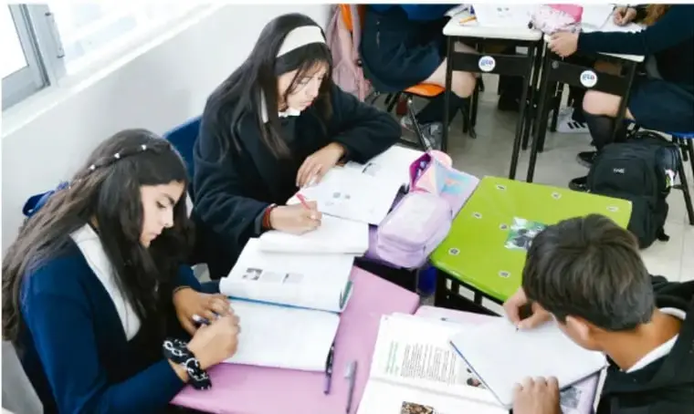 Fotografía de estudiantes de telesecundaria trabajando en grupo alrededor de mesas con cuadernos y materiales escolares, dialogando y escribiendo.