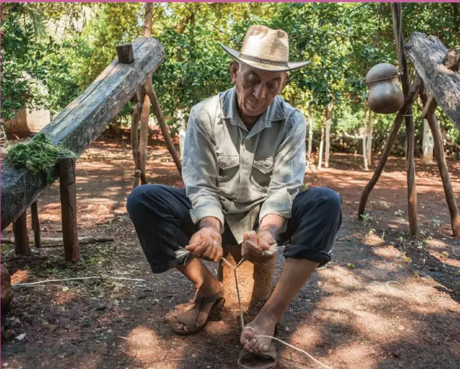 Fotografía a color de un hombre adulto mayor con sombrero de paja, camisa y pantalón oscuro, sentado en un banco de madera al aire libre; torsiona con las manos fibras o hilos largos (tejido artesanal). Alrededor hay troncos, un recipiente de barro colgado de una horqueta y vegetación verde.