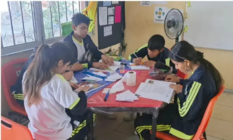 Fotografía de cuatro estudiantes de telesecundaria con uniforme trabajando en equipo alrededor de una mesa forrada con papel, con recortes, periódicos y materiales de plástico; aula con ventana, ventilador y pizarrón al fondo.