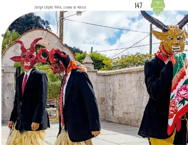 Dos hombres con máscaras rojas de diablos y trajes oscuros, uno con falda de fibra natural, caminando en un patio con muro de piedra.
