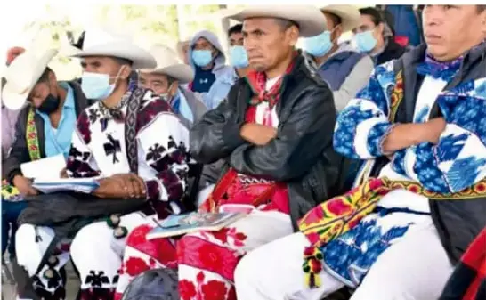 Grupo de personas indígenas con vestimenta tradicional, tocados y pintura corporal, portando bastones y objetos ceremoniales en un paisaje árido.