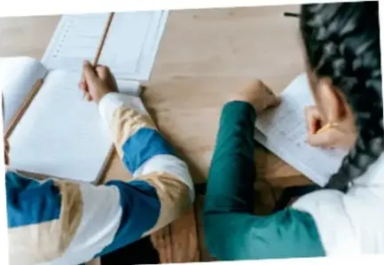 Fotografía de dos estudiantes escribiendo en sus cuadernos sobre una mesa de madera.