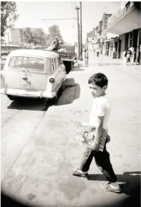 Fotografía en blanco y negro de un niño caminando por una calle con un libro, junto a un automóvil antiguo.