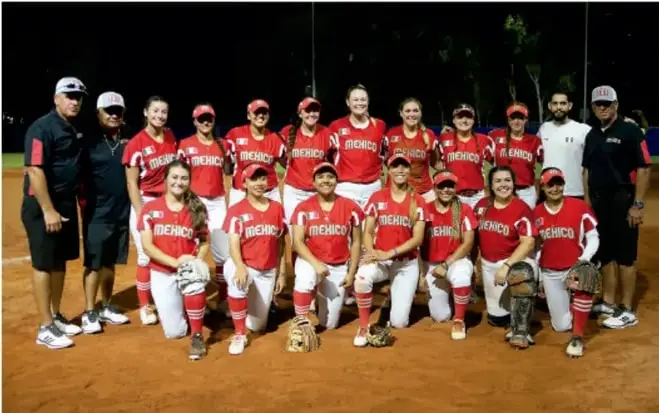 Fotografía de un equipo de softball o béisbol femenil de México: jugadoras con uniforme rojo con la palabra MÉXICO, junto con cuerpo técnico, posando en el campo de juego durante la noche.