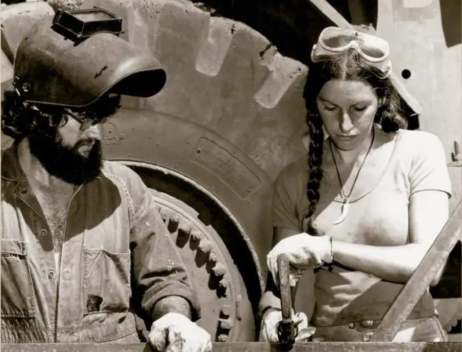 Fotografía histórica en blanco y negro titulada 'Mecánica', ca. 1980: un hombre con casco de soldador y una mujer con gafas trabajando juntos en una pieza mecánica grande. Acervo INEHRM.