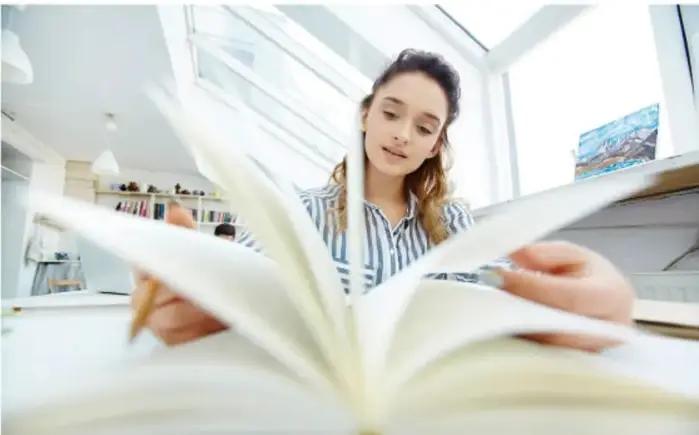 Fotografía de una estudiante leyendo un libro abierto en un espacio amplio y luminoso, frente a una ventana.