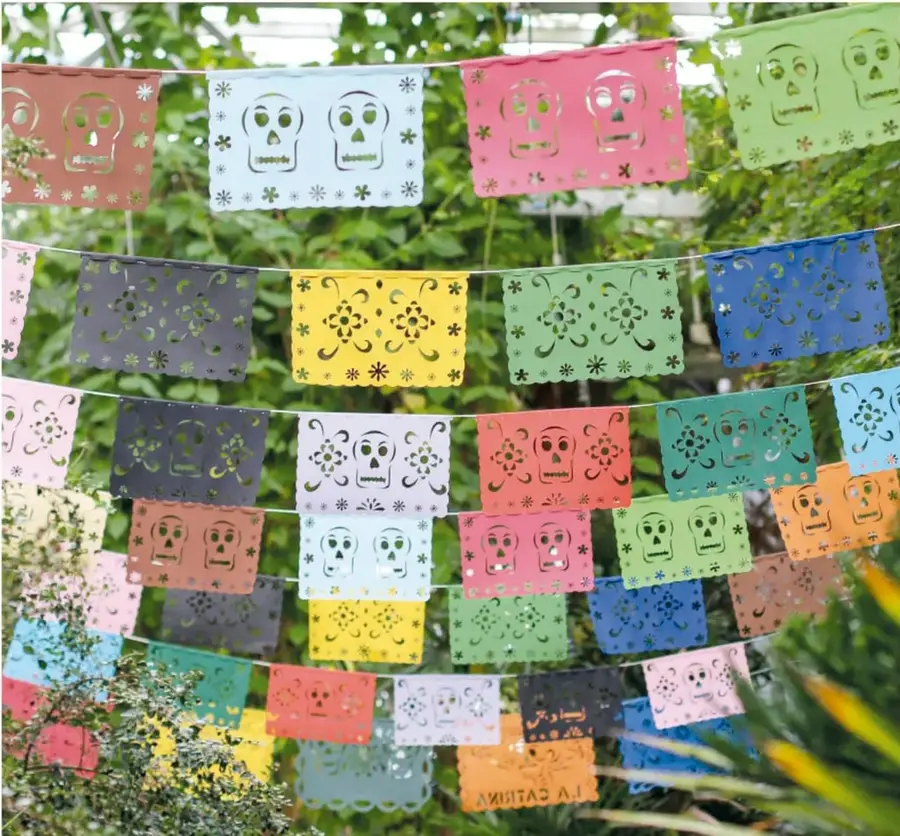 Hileras de banderolas de papel picado multicolor con calaveritas y flores caladas, colgadas al aire libre entre vegetación.