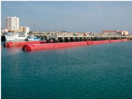 Fotografía de un atenuador de superficie: una estructura larga roja flotando sobre el agua, conformada por varias partes articuladas, en una zona costera con barcos y edificios al fondo.