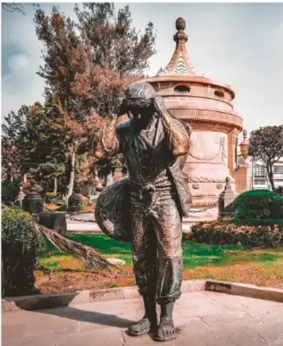 Fotografía a color de la escultura El Aguador: figura de bronce de un hombre que carga a la espalda una gran vasija de barro, junto a un árbol, con la Caja del Agua (estructura circular de cantera con techo cónico) al fondo entre jardines.