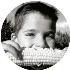 Fotografía en blanco y negro de un niño sonriente sosteniendo una mazorca de maíz cerca de su boca.