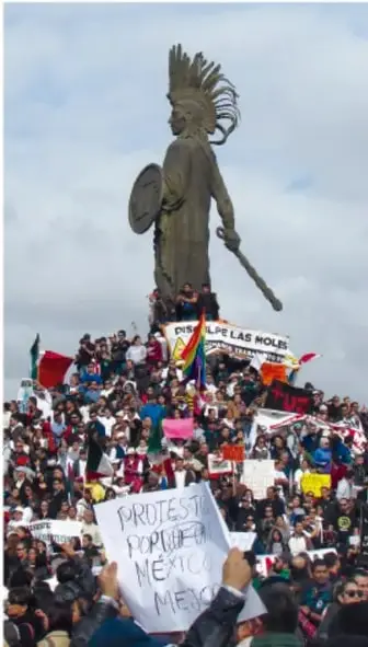 Monumento a una figura indígena con tocado, rodeado de una multitud con pancartas que dicen 'Protesta por México' y 'Respeten las raíces'.