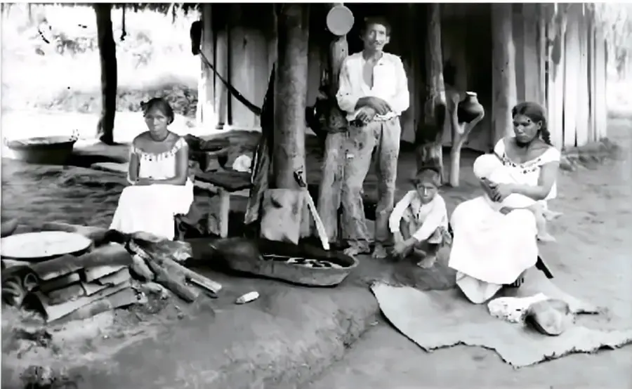 Fotografía histórica en blanco y negro de una familia indígena junto a una vivienda de palapas, con mujeres sentadas, un hombre de pie y niños.