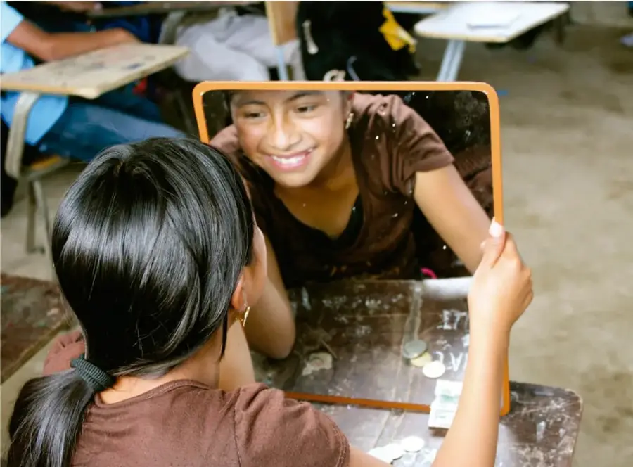 Fotografía de una estudiante de secundaria de espaldas, con cabello recogido en coleta, mirándose sonriente en un espejo pequeño de marco naranja que sostiene con ambas manos sobre su pupitre de madera en el aula.