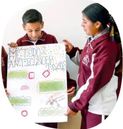 Fotografía de dos estudiantes de telesecundaria con uniforme (chico y chica) sosteniendo un cartel elaborado por ellos que dice "MÉTODOS ANTICONCEPTIVOS" con dibujos y recortes de colores.