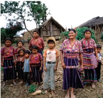 Fotografía de un grupo de personas indígenas, en su mayoría mujeres y niños con vestimenta tradicional, de pie frente a una vivienda rural de madera y techo de lámina, en un entorno natural.