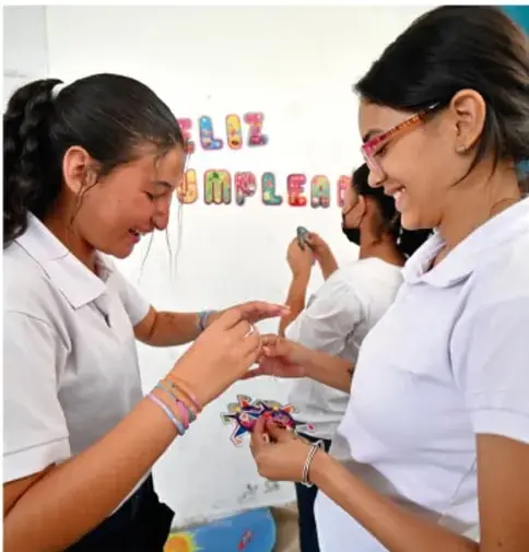 Fotografía de dos estudiantes mujeres sonriendo e intercambiando pulseras de colores, en un ambiente escolar. Al fondo se ve un cartel con la palabra 'EMPLEA' y otras letras.