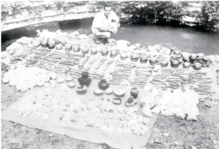 Fotografía en blanco y negro de una mesa o superficie al aire libre cubierta con numerosos objetos arqueológicos pequeños (vasijas, figurillas, cuentas) dispuestos en hileras; al fondo se observa una persona sentada junto a un cuerpo de agua, probablemente el cenote.