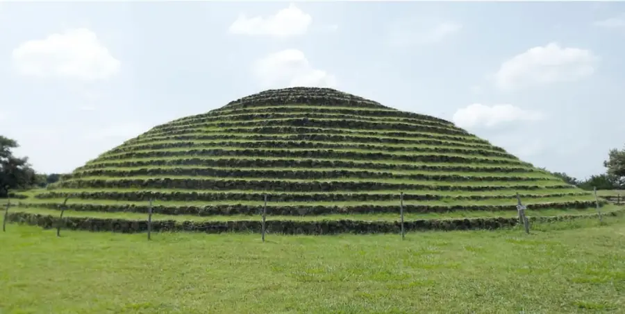 Fotografía frontal de una gran pirámide circular con múltiples anillos escalonados cubiertos de césped, rodeada de pasto y una cerca.