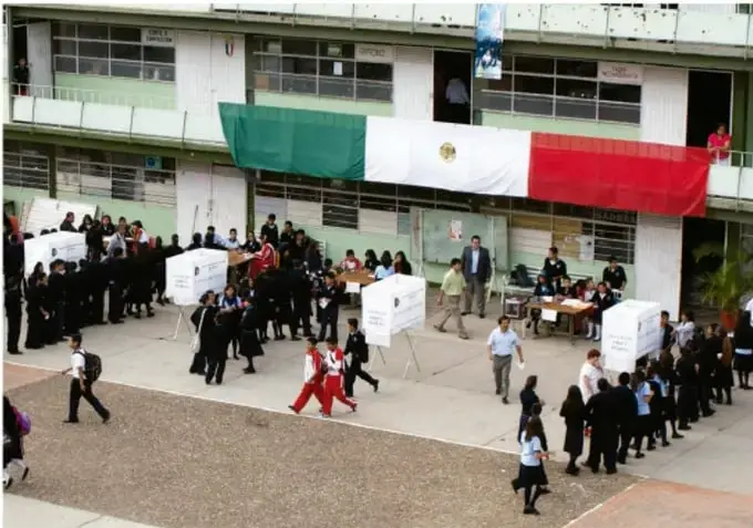 Fotografía de una escuela con la bandera de México desplegada en el patio y alumnos formados; parece una actividad cívica o simulación de votación, lo que ilustra la participación democrática escolar.