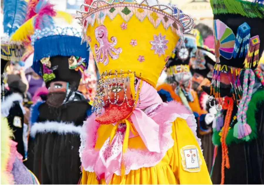 Fotografía de Carlos Hahn (Ciudad de México) que muestra a varios chinelos en comparsa: un chinelo con traje amarillo y rosa y máscara con barba en primer plano, y otros con trajes y sombreros ornamentados de colores vivos (azul, verde, negro).