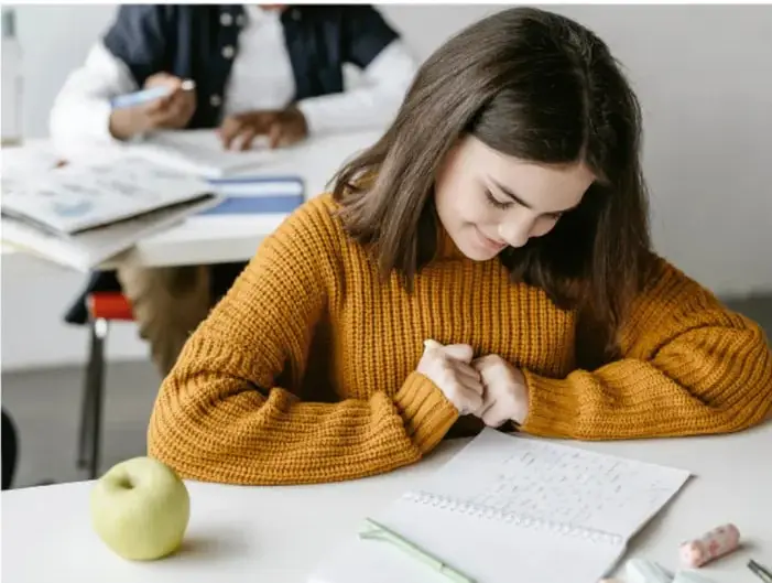 Fotografía de una estudiante con suéter amarillo escribiendo en su cuaderno en un salón de clases; al fondo se ve otro estudiante y una manzana verde sobre la mesa.