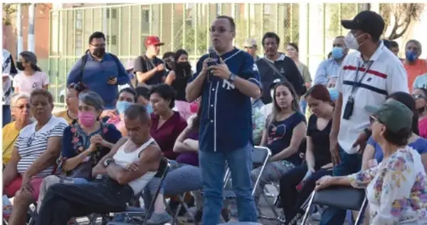 Fotografía de una reunión comunitaria al aire libre: una persona habla con un micrófono frente a un grupo de vecinos sentados, algunos con cubrebocas, en un espacio público.