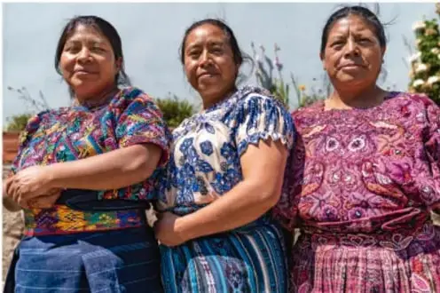Tres mujeres mayas con trajes tradicionales de colores vivos, de pie y sonriendo.