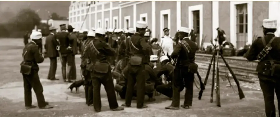 Fotografía histórica en sepia de soldados federales armados con fusiles frente al edificio de la fábrica de hilados de Río Blanco, Veracruz, 1907; se observan vías de tren, civiles y un perro.
