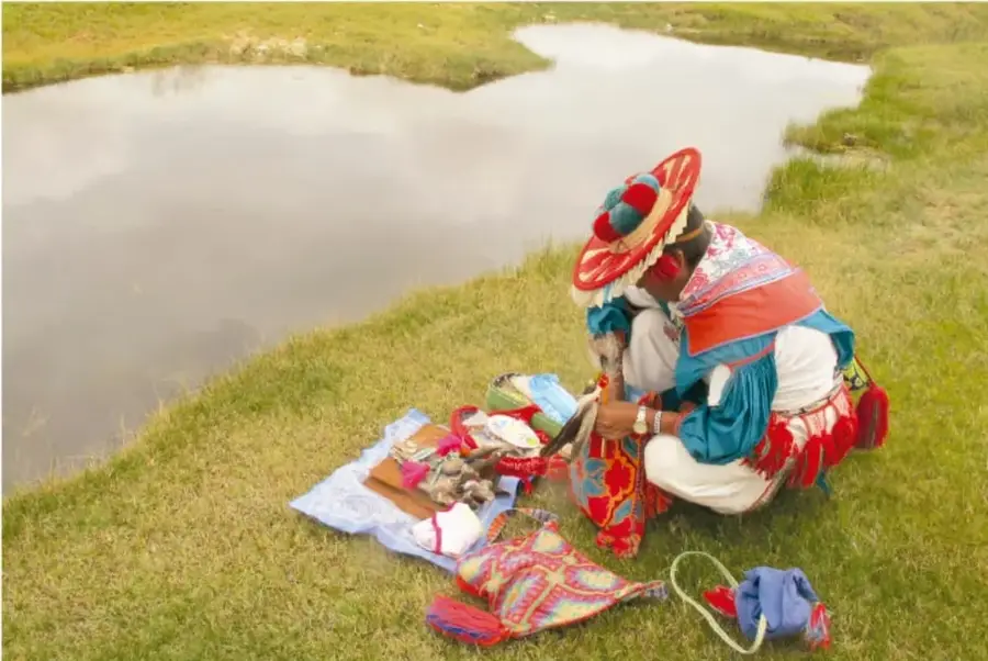 Fotografía de un peregrino wixaritari con sombrero tradicional de colores, sentado junto a un estanque de agua, preparando ofrendas sobre telas bordadas.