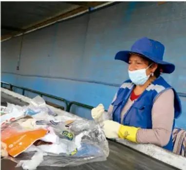 Fotografía de una trabajadora con casco, mascarilla y guantes clasificando residuos sólidos en un sitio de tratamiento de desechos, ilustrando la gestión de productos secundarios de la comunidad.
