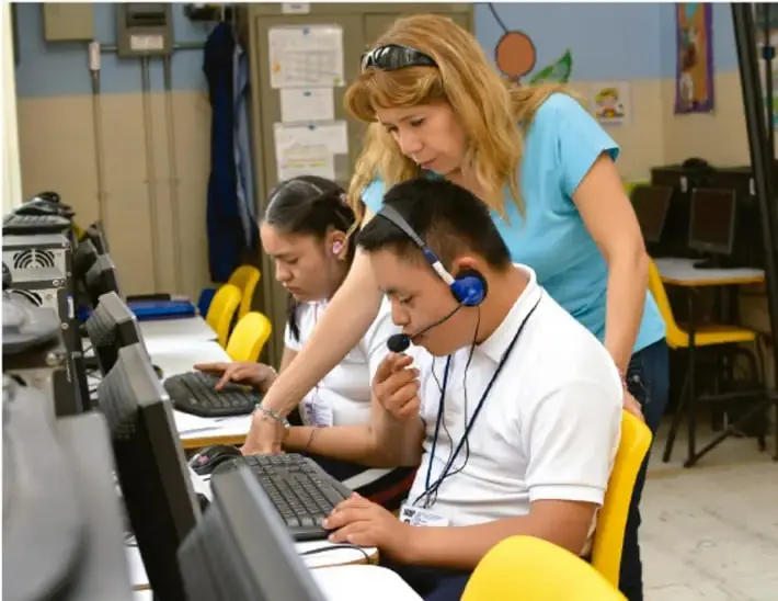 Fotografía de estudiantes en un aula de cómputo con audífonos, trabajando en computadoras mientras una docente los orienta; ilustra el uso de formatos digitales para presentar hallazgos del proyecto.