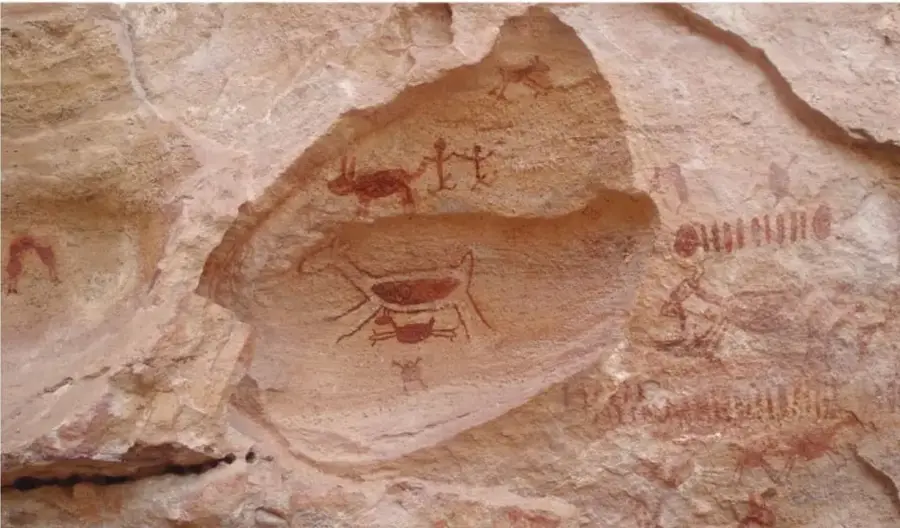 Fotografía de pinturas rupestres sobre roca, con figuras humanas y animales trazadas en pigmentos rojos y ocres sobre una pared de piedra clara.