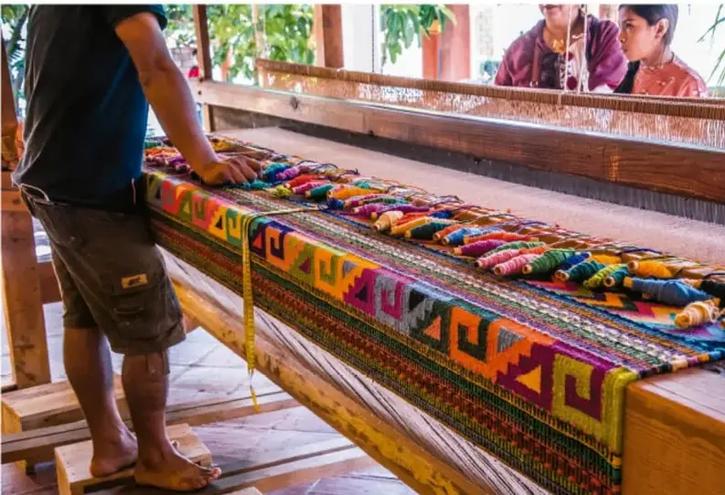 Fotografía de personas trabajando en un telar de gran formato, tejiendo textiles de colores con hilos dispuestos sobre el aparato; representa una actividad productiva artesanal en la comunidad.