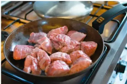 Fotografía de trozos de carne cocinándose en un sartén sobre una estufa, ilustrando la conducción de calor.