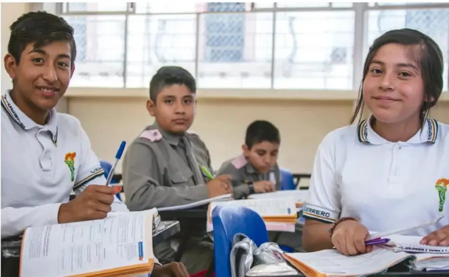 Cuatro estudiantes adolescentes en un salón de clases con uniformes blancos, trabajando en sus cuadernos y libros; una chica y un chico sonríen a la cámara.