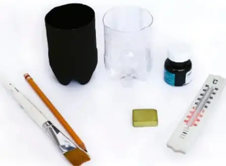 Fotografía de los materiales del experimento: dos botellas cortadas (una pintada de negro y una transparente), un pote de pintura negra, una brocha, dos termómetros de laboratorio, un borrador y un lápiz sobre una superficie blanca.