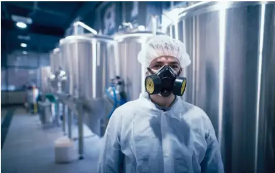 Trabajador industrial con overol blanco, cofia y respirador con filtros amarillos, de pie frente a tanques metálicos inoxidables en una planta de proceso.