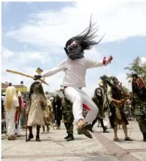 Fotografía de una danza o manifestación cultural comunitaria: un participante con máscara y cabello al aire baila en la calle junto a otros asistentes, en una celebración tradicional.